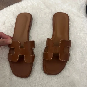 H tofflor - Snygga bruna sandaler med breda remmar och dekorativ vit söm. De har fyrkantig tå och platt sula, perfekt för en chill sommarlook. Tillverkade i skinn för en lyxig känsla och passar dig som gillar stilrena detaljer.