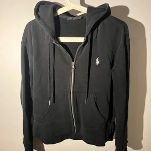 Svart hoodie från Polo Ralph Lauren - Svart hoodie från Polo Ralph Lauren med dragkedja och huva. Klassisk design med vit broderad logga på bröstet, ( en dam tröja ) 