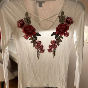 Vit långärmad topp med broderade blommor - Säljer en vit långärmad topp från Amisu i storlek XS. Toppen har snygga broderade röda och rosa blommor framtill, v-ringning med korsade band och är gjord i tunn bomull. Perfekt för dig som gillar romantiska detaljer och vill sticka ut lite extra.