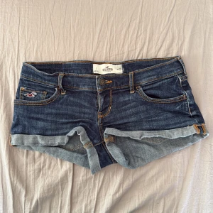 Mörkblå jeansshorts från Hollister - Snygga mörkblå jeansshorts från Hollister med klassisk femficksdesign och uppvikta ben. Shortsen har kontrastsömmar i gult och detaljerad brodering på bakfickorna. Perfekt för varma dagar och har en lågmidjad passform.