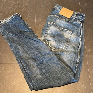 Jeans med slitningar - Säljer ett par blå jeans med snygga slitningar och ljusa tvättade partier. Klassisk femficksmodell med röd knappdetalj och tydliga sömmar på bakfickorna. Jeansen har en smal passform och är tillverkade i jeansmaterial.