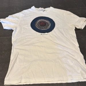 Vit t-shirt med tryck från Ben Sherman - Vit t-shirt från Ben Sherman med ett stort, cirkulärt tryck i blått, rött och svart på bröstet. Klassisk rund hals och korta ärmar. Materialet är mjuk bomull som känns skönt mot huden. Perfekt för dig som gillar grafiska prints och stilren design.