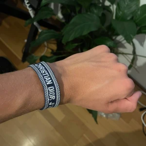 Blått Christian Dior J'Adior armband - Snyggt blått tygarmband från Christian Dior med texten 'J'Adior' broderad i vitt. Armbandet har en randig design med vita detaljer längs kanterna och är justerbart med knytning. Perfekt för dig som vill ha en cool designeraccessoar på handleden.