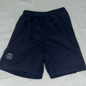 PSG mörkblå träningsshorts - Mörkblå träningsshorts från Paris Saint-Germain med klubbens emblem på vänster ben. Klassisk sportig look med elastisk midja och tillverkade i lätt polyester som andas. Perfekta för fotbollsträning eller gymmet.