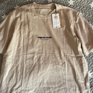 Beige t-shirt från Jack & Jones - Snygg beige t-shirt från Jack & Jones Originals Studio med svart tryck på bröstet. Klassisk rund hals och korta ärmar. Mjuk och skön bomull som passar perfekt till jeans eller shorts. Enkel och clean design med modern vibe.