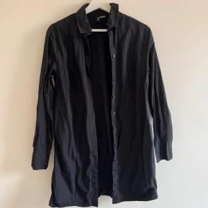 Svart skjortklänning - Svart lång skjortklänning från H&M Divided i storlek XS. Skjortan har klassisk krage, långa ärmar med manschett och knappar hela vägen framtill. Tillverkad i mjuk bomull, perfekt för en avslappnad och stilren look.Varsamt använd, ser inga skavanker. Jag har massor av artiklar till salu! Köp flera och få rabatt!