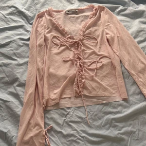 Ljusrosa blus med snörning från Weekday - Supersöt ljusrosa blus från Weekday med långa ärmar, volangkant längs v-ringningen och snörning framtill. Tunn och luftig i bomullsmix, perfekt för vår och sommar. Ger en romantisk vibe med sina fina detaljer och feminina form.