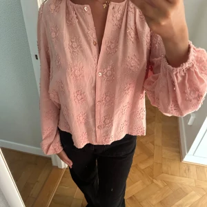 Rosa blommig skjorta/blus från Dobber - Supersöt rosa skjorta/blus från Dobber med broderade blommor över hela tyget. Skjortan har långärmade puffärmar med resår vid ärmslut, knappar framtill och en loose passform.