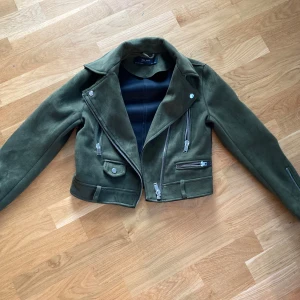 Olivgrön mockajacka från Zara  - Snygg olivgrön mockajacka från Zara med cool bikerstil. Jackan har flera silvriga dragkedjor, snedställda fickor och klassisk krage. Croppad modell som ger en edgy vibe, perfekt till jeans eller klänning. Materialet är mjuk mocka och jackan har långa ärmar med dragkedjor vid ärmsluten.