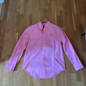 Rosa skjorta från Polo Ralph Lauren - Snygg rosa skjorta från Polo Ralph Lauren i bomull med blå broderad logga på bröstet. Skjortan har lång ärm, klassisk krage med knappar och slim fit-passform. Perfekt för dig som gillar stilrena och färgglada plagg.