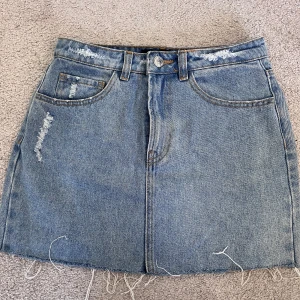 Blå jeanskjol från BikBok med slitningar - Säljer en snygg blå jeanskjol från BikBok i storlek 36. Kjolen är kort med råa kanter och slitna detaljer både fram och bak. Klassisk femficksmodell med knapp och dragkedja framtill. Perfekt för dig som gillar en avslappnad och trendig stil.