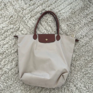 Beige shoppingväska från Longchamp - Snygg beige shoppingväska från Longchamp med bruna skinnhandtag och lock. Har inte använt mycket men är lite smutsig på kanten men är perfekt storlek för skolstarten. Böcker mappar och dator+ extra får plats i väskan till skolan 