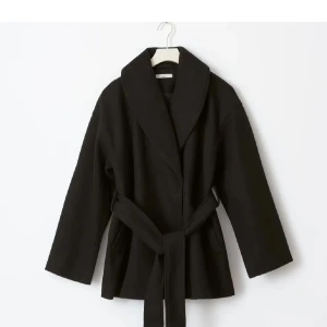 Svart omlottkappa med bälte - Stilren svart kappa med bred krage och knytbälte i midjan. Jackan har en avslappnad passform och långa ärmar, perfekt för kyligare dagar. Enkel och snygg design som passar till många outfits.