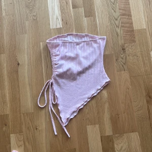 Ljusrosa ribbad tubtopp med snörning - Supersöt ljusrosa tubtopp i bandeau-stil med ribbad struktur och snörning i sidan. Toppens asymmetriska form ger en trendig look och den är perfekt för varma dagar. Mjuk och stretchig, lätt att styla till jeans eller kjol.