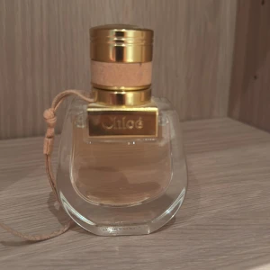 Chloé Nomade Eau de Parfum - Chloé Nomade Eau de Parfum i en elegant, genomskinlig glasflaska med guldiga detaljer och beige mockaband runt halsen. Flaskan har en mjukt rundad form och exklusiv känsla med märkets logga i guld framtill. Doften är ljus bärnstensfärgad.