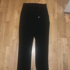 Svarta högmidjade kostymbyxor - Snygga svarta kostymbyxor med hög midja och dekorativa knappar framtill. Byxorna har raka ben och en stilren look som passar perfekt till en dressad outfit. Materialet känns mjukt och följsamt, vilket gör dem bekväma att bära hela dagen.