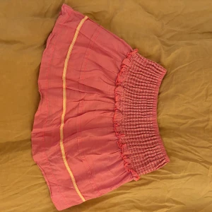 Rosa volangkjol med smock - Söt rosa kortkjol med volang och smockad midja. Kjolen har ljusgula dekorativa sömmar och är gjord i ett lätt bomullstyg som ger en skön och luftig känsla. Perfekt för dig som gillar färg och vill ha en feminin look.