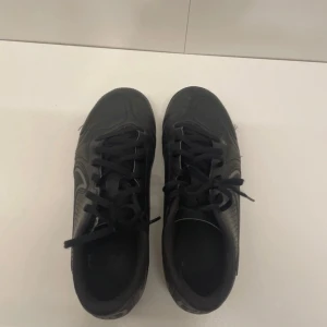 Nike Tiempo svarta fotbollsskor - Svarta Nike Tiempo fotbollsskor med klassisk design och snörning. Skorna har rund tå, platt sula med fasta dobbar och är tillverkade i skinn med syntetdetaljer. Swoosh-logga på sidan och Tiempo-tryck bak. Perfekta för dig som vill ha stilrena och funktionella skor på planen. Dom är använda men i bra skick. 