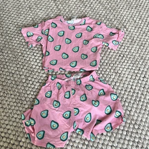  Rosa pyjamasset med avokado mönster - Säljer ett pyjamasset med rosa shorts och tröja. Shortsen har elastisk midja och ett lekfullt avokadomönster i grönt och gult. Materialet är mjuk bomull, perfekt för häng på sommaren. Passformen är loose och de har en relaxed vibe. Tröjan har samma avokadomönster som shortsen och har en loose och lite kortare passform.