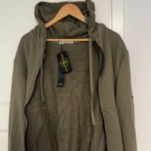 Stone Island kofta - Snygg olivgrön/khali kofta från Stone Island med dragkedja och huva. Jackan har klassisk logotyp på ärmen, snörning i huvan och är tillverkad i mjuk bomull. Perfekt för dig som gillar streetwear och vill ha en stilren look.