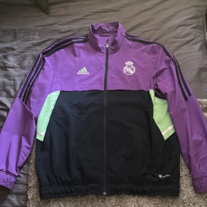 Real Madrid vindjacka/tröja Adidas L - Snygg vindjacka från Adidas x Real Madrid i lila och svart med gröna detaljer. Jackan har dragkedja, höga krage och broderade loggor på bröstet. Tillverkad i lätt polyester, perfekt för fotboll eller chill. Elastiska muddar och klassiska Adidas-ränder på axlarna. Köpt på unisport!