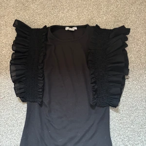 Svart topp med volangärmar från Amisu - Svart topp från Amisu i storlek XS med unika volangärmar i transparent tyg och smockdetaljer. T-shirten har rund halsringning och är tillverkad i mjukt material. Perfekt för dig som vill sticka ut med coola detaljer och volym på axlarna.