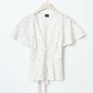 Blommig omlottopp från Gina Tricot, S - Supersöt vit omlottopp från Gina Tricot med ljusblått blommönster. Toppen har korta ärmar, v-ringning och knytband i midjan. Säljer då den är för stor. Har inte använt den bara provat 💕