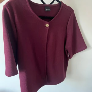 Vinröd cardigan jacka från Gina Tricot - Snygg vinröd cardigan från Gina Tricot med rund halsringning och en guldig knapp framtill. Koftan har trekvartsärm och är gjord i ett mjukt material.