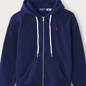 Marinblå hoodie från Polo Ralph Lauren - Snygg marinblå hoodie från Polo Ralph Lauren med vit snörning och dragkedja framtill. Klassisk röd logga på bröstet och sköna fickor. Perfekt för chill dagar och enkel att matcha med jeans eller joggers. Ord pris 2145