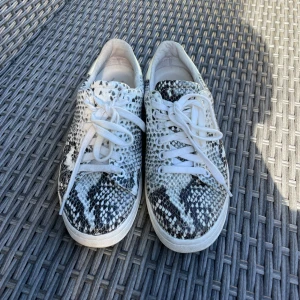 Sneakers med ormmönster från K. Cobler - Snygga sneakers från W. Cobler med vit bas och svart ormmönster. Skorna har vita snören, rund tå och platt sula. Ovandelen är i syntetmaterial med reptilstruktur och detaljer i blankt vitt bak. Perfekta för dig som vill sticka ut med coola detaljer.Orm reptil mönstrade sneakers skor från Kcobler. Använda men i bra skick, vg se bilder. Äkta skinn läder. Storlek 36, normal i storlek.