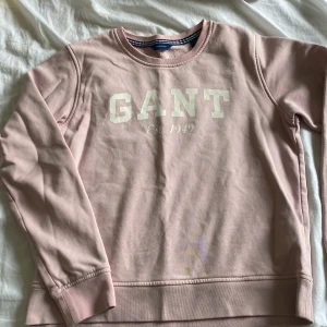 Rosa sweatshirt från GANT - Säljer en ljusrosa sweatshirt från GANT med vit logotyp och text 'Est. 1949' på bröstet. Tröjan har rund hals, långa ärmar och ribbade muddar. Mjuk insida i fleece och klassisk passform. Perfekt för chill dagar och enkel att matcha med jeans eller leggings.