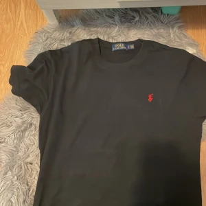 Äkta svart Polo raplh Lauren T Hsirt Round neck - Svart t-shirt från Polo Ralph Lauren med klassisk röd broderad logga på bröstet. Modellen är kortärmad och har en rund halsringning. Materialet är mjuk bomull som känns skönt mot huden. Perfekt för dig som gillar stilrena och ikoniska plagg.