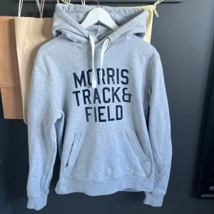 Grå hoodie Morris - Snygg grå hoodie från Morris Gents med trycket 'Morris Track & Field' i mörkblått på bröstet. Tröjan har huva med vita snören och en klassisk känguruficka framtill. Perfekt för chill dagar och sportiga vibes. Hoodien är väldigt svår att få tag i.