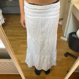 Vit långkjol med broderi från Gina Tricot - Supersöt vit långkjol från Gina Tricot med broderade blommor och smock i midjan. Kjolen är gjord i mjuk bomull och har volang nertill för extra flow. Perfekt för sommarens alla häng och picknickar. Passar dig som gillar romantisk och chill stil.