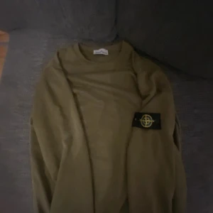 Olivgrön Stone Island sweatshirt - Säljer en olivgrön sweatshirt från Stone Island med klassisk patch på vänster ärm. Tröjan har rund halsringning och är tillverkad i mjuk bomull. Perfekt för dig som gillar streetwear och vill ha en clean look med ikonisk detalj.
