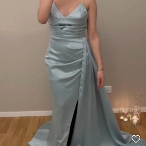 Ljusblå långklänning med slits - Säljer en elegant ljusblå långklänning med smala axelband och v-ringad urringning. Klänningen har en snygg drapering i midjan och en hög slits framtill som ger en cool touch. Materialet är glansigt och känns lyxigt, perfekt för bal eller nyår. En till med en ny storlek