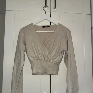 Beige omlott tröja från Bik Bok - Säljer en beige omlott tröja från Bik Bok med croppad passform och bred ribbad midja. Tröjan har långärmade ärmar och djup v-ringning framtill. Mjuk och stretchig i bomullsmix, perfekt för chill dagar eller när du vill vara extra comfy. Sömmen har gått upp se på bilden. 