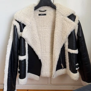 trendig teddy bikerjacka från Gina Tricot - Säljer en svart teddyjacka från Gina Tricot med feta vita teddy-detaljer och foder. Jackan har en oversized krage, dragkedja och coola kontraster mellan det svarta skinnet och det fluffiga vita. Perfekt för kalla dagar när du vill sticka ut.