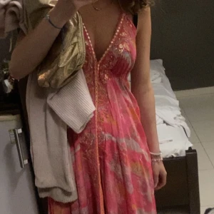 Rosa långklänning med blommigt mönster - Säljer en superfin långklänning i rosa och orange toner med blommigt mönster och broderade detaljer. Djup v-ringning och ärmlös modell, perfekt för sommarens alla tillfällen. Klänningen har en flowy passform och vackra detaljer längs urringningen. Använt den en gång!