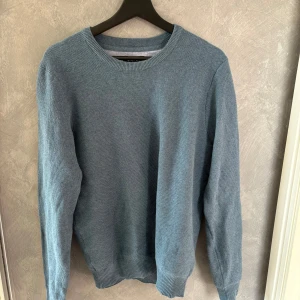 Blå stickad tröja med rund hals - En stilren blå stickad tröja med rund halsringning och ribbade muddar vid ärmslut och nederkant. Perfekt för lager-på-lager och enkel att matcha med jeans eller chinos. Långärmad och i mjukt material som känns skönt mot huden.