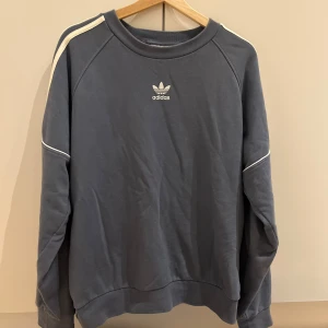 Blå Adidas sweatshirt med vita detaljer - Snygg blå set från Adidas med klassiska vita ränder längs ärmarna och broderad logga på bröstet. Tröjan har rund hals, lång ärm och är gjord i mjuk bomullsmix. Perfekt för en sportig och avslappnad stil.