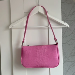 Rosa axelväska i krokopräglat skinn - Säljer en  rosa axelväska med krokopräglat mönster och silverfärgad dragkedja. Väskan har axelband. Perfekt för att lyfta vilken outfit som helst.