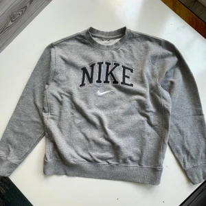 Nike sweatshirt - Snygg grå sweatshirt från Nike med stort svart broderat NIKE-tryck och vit swoosh på bröstet. Klassisk rund halsringning och ribbade muddar vid ärmslut och nederkant. Perfekt för en chill och sportig look.