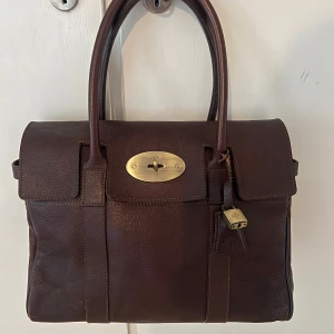 Mulberry Bayswater brun handväska skinn - Ej äkta, köpt på second hand. Klassisk Mulberry Bayswater handväska i mörkbrunt skinn med snygga mässingsdetaljer och lås. Väskan har två rejäla handtag, ikonisk vridknapp framtill och präglad Mulberry-logga. Perfekt storlek för vardagsgrejer och stilren look.