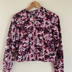 Blommig vindjacka från stadium  - Snygg vindjacka från XTTL med blommigt mönster i rosa, lila och vitt. Jackan har dragkedja framtill, långärmade ärmar och en lätt, sportig känsla. Perfekt för dig som gillar färg, passar bra till idrott.