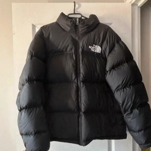 Svart pufferjacka från The North Face - Säljer en svart pufferjacka från The North Face med klassisk logga på bröstet och ryggen. Jackan har hög krage, dragkedja framtill och är riktigt fluffig. Perfekt för kalla dagar när du vill hålla stilen och värmen. Passformen är normal och jackan är dunfylld. Storlek L i barnstorlek men passar som xs!❤️❤️