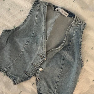 Ljusblå jeansväst från Zara - Säljer en snygg ljusblå jeansväst från Zara med klassisk v-ringning och knappar framtill. Västen har en rak passform och är tillverkad i denim med tydliga sömmar. Aldrig använd!
