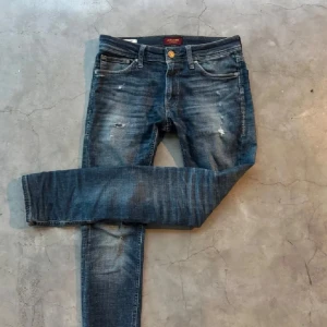 Blå slim jeans från Jack & Jones - Snygga blå slim  jeans från Jack & Jones med slitningar och tvättade detaljer. Klassisk femficksmodell med gylf och knapp, samt läderpatch bak i midjan. Perfekta för en avslappnad och trendig look.