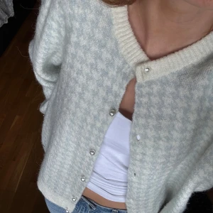 Ljusblå och vit kofta med pärlknappar - Superfin kofta i ljusblått och vitt med diskret hundtandsmönster. Rund halsringning, ribbade muddar och pärlknappar framtill. Mjuk och fluffig känsla, perfekt till jeans eller kjol. Långärmad och i stickad ullmix för extra mysfaktor.