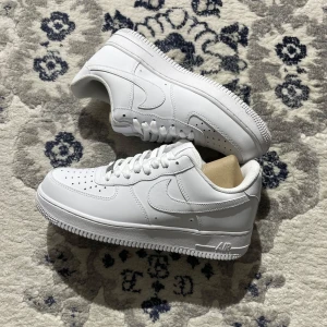 Nike Air Force 1 - Klassiska Nike Air Force 1 sneakers i helvitt skinn med perforerad tå, rund tå och platt sula. Snygg och tidlös modell med snörning och ikonisk swoosh-logga på sidorna. Perfekta för dig som vill ha en clean och stilren look.